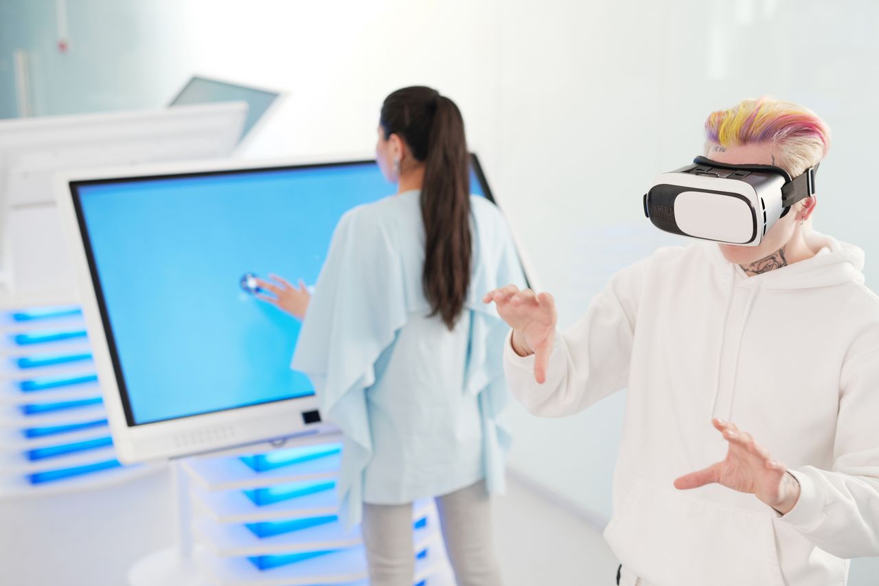 services-01 Two young adults engaging with VR and touchscreen technology in a futuristic room.